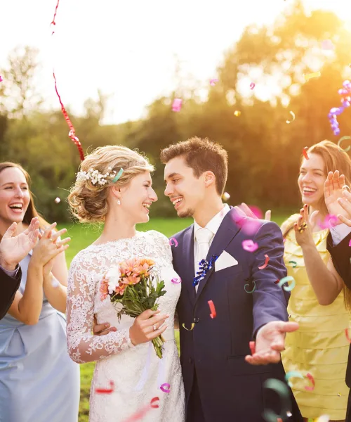 Full length portrait of newlywed couple and their friends at the wedding party showered with confetti in green sunny park